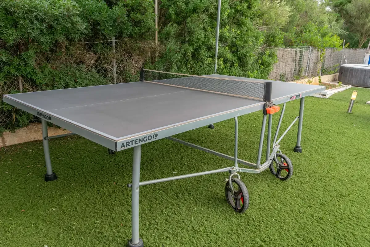 An outdoor table tennis table stands on a lawn beside dense greenery.