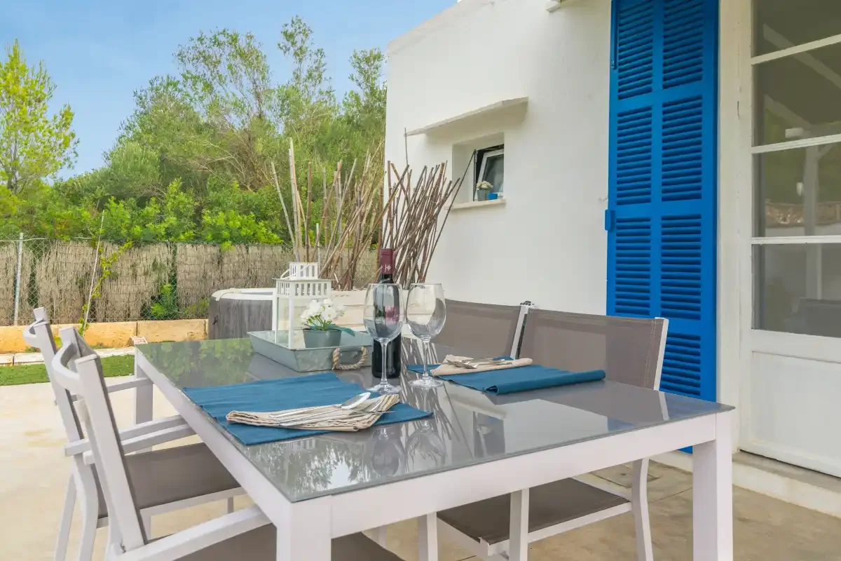 An outdoor dining area beside a white building, with a glass-topped table and blue shutters.