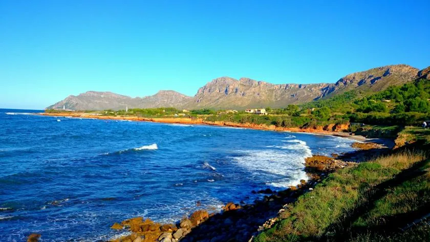A rugged coastline curves beside blue water, with green hills in the distance.