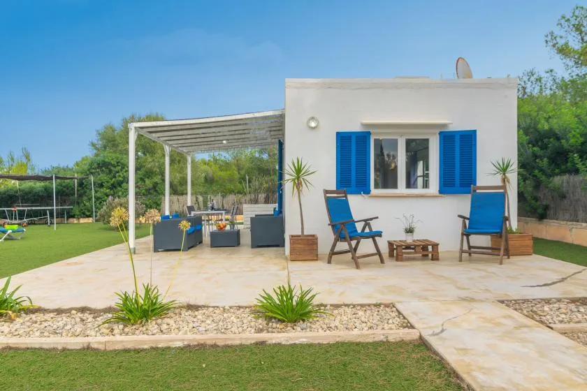 A small white house with blue shutters opens onto a sunny patio and pergola.