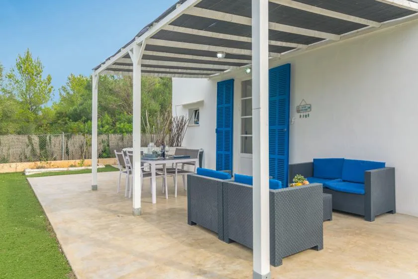 A covered patio has outdoor seating beside bright blue shutters and a green lawn.