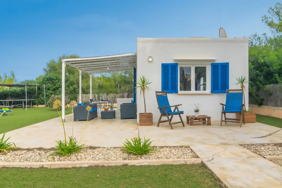A white house with blue shutters opens onto a paved patio with outdoor seating.