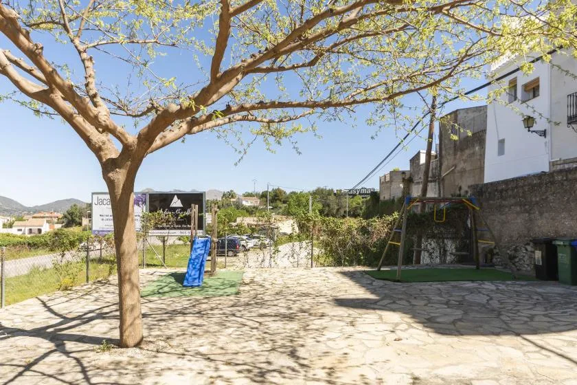 Zona de juegos al aire libre bajo un árbol frondoso, con tobogán y estructura para trepar.