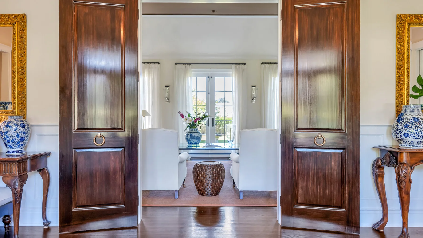 Una sala luminosa se ve a través de puertas de madera abiertas, con un jarrón sobre una mesa.