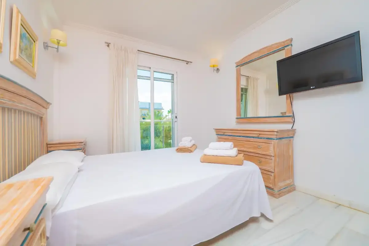 Un dormitorio luminoso con una cama tendida, una cómoda y un televisor en la pared.