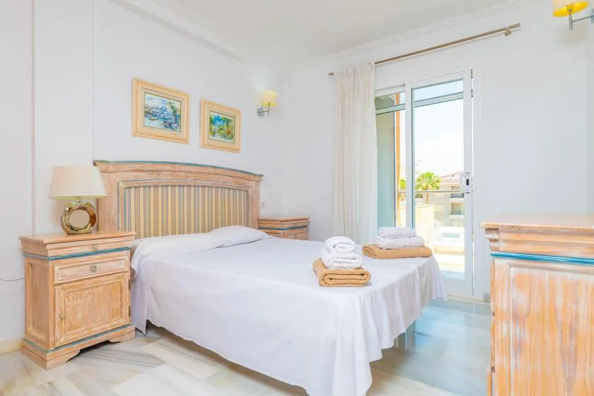 Dormitorio con una cama blanca, muebles de madera y una puerta acristalada que da a un balcón.