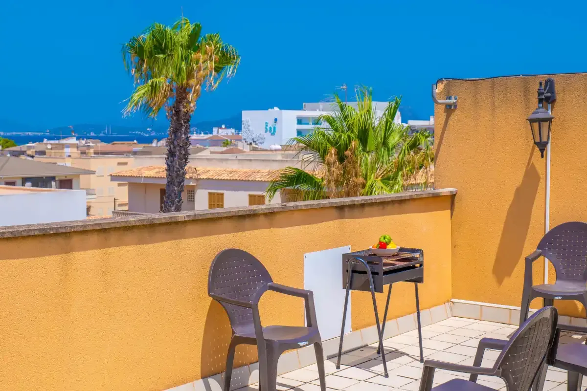 Una terraza soleada tiene asientos y una mesa pequeña, con palmeras y edificios al fondo.