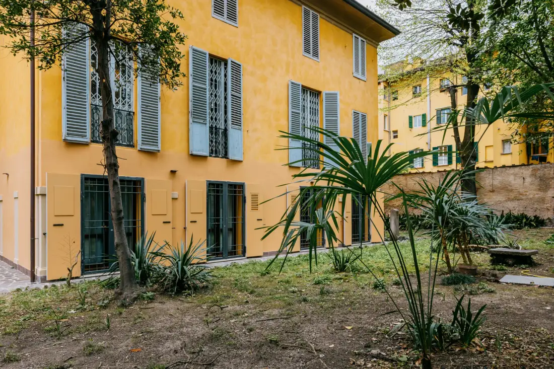 Un edificio amarillo con ventanas con persianas se alza detrás de un pequeño jardín.