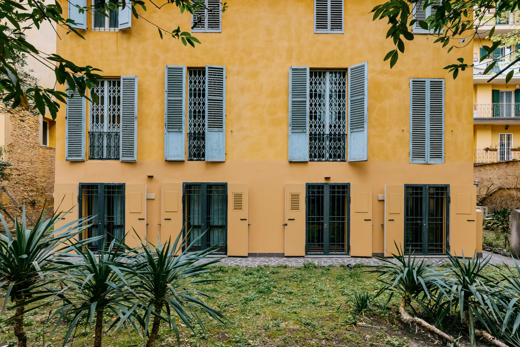 Una fachada amarilla con ventanas con contraventanas da a un pequeño jardín.