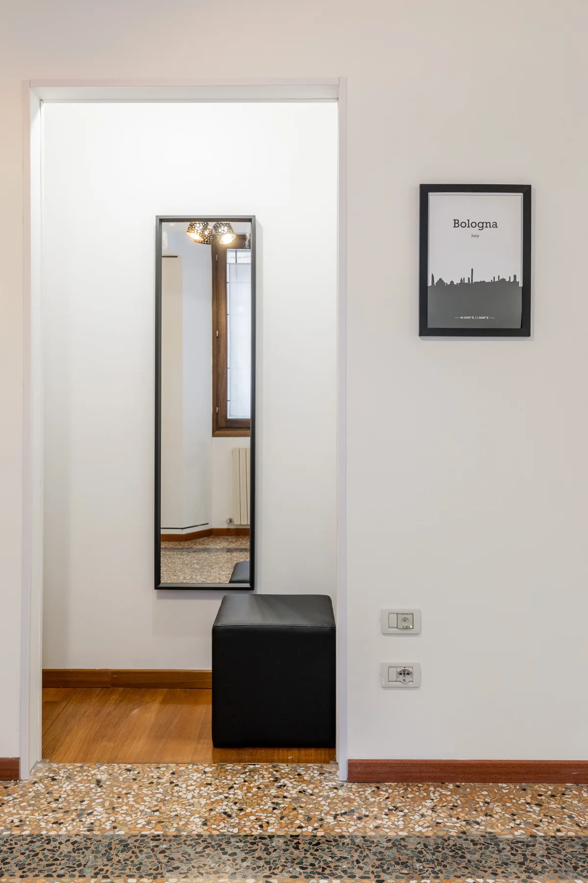 Un pasillo luminoso con un espejo alto, un asiento oscuro y un cuadro enmarcado.