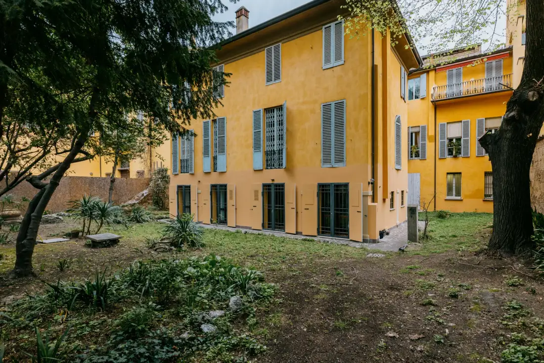 Un edificio amarillo de varias plantas con ventanas con contraventanas junto a un jardín.