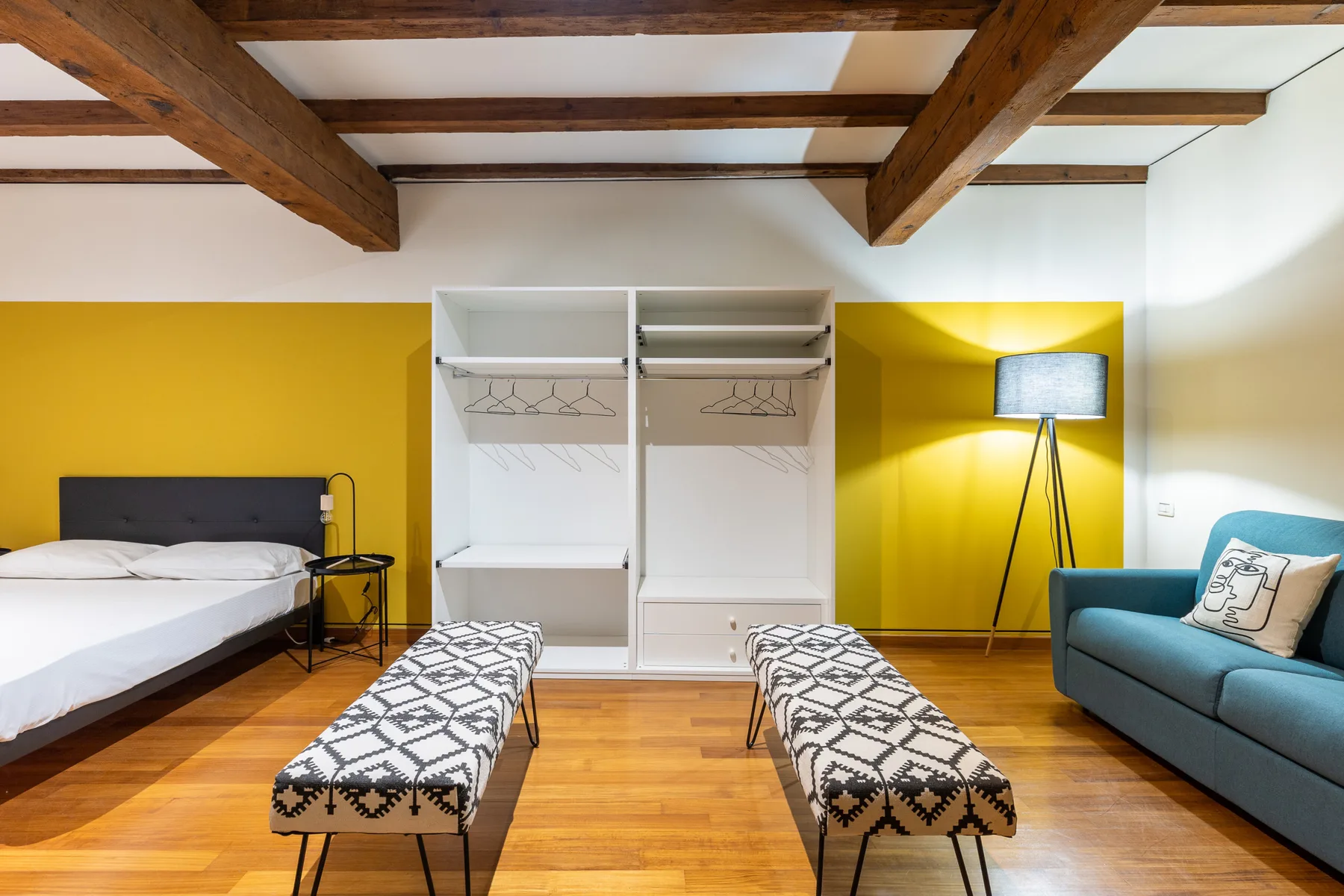 Un dormitorio con una cama, dos bancos estampados y vigas de madera vistas en el techo.