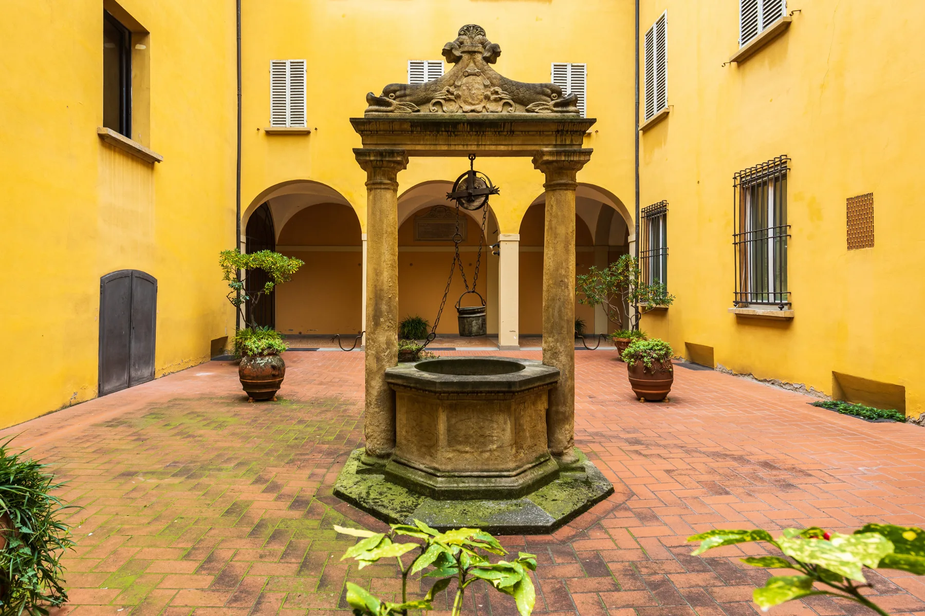 Un patio de ladrillo tiene un pozo de piedra ornamentado y pasillos con arcos al fondo.