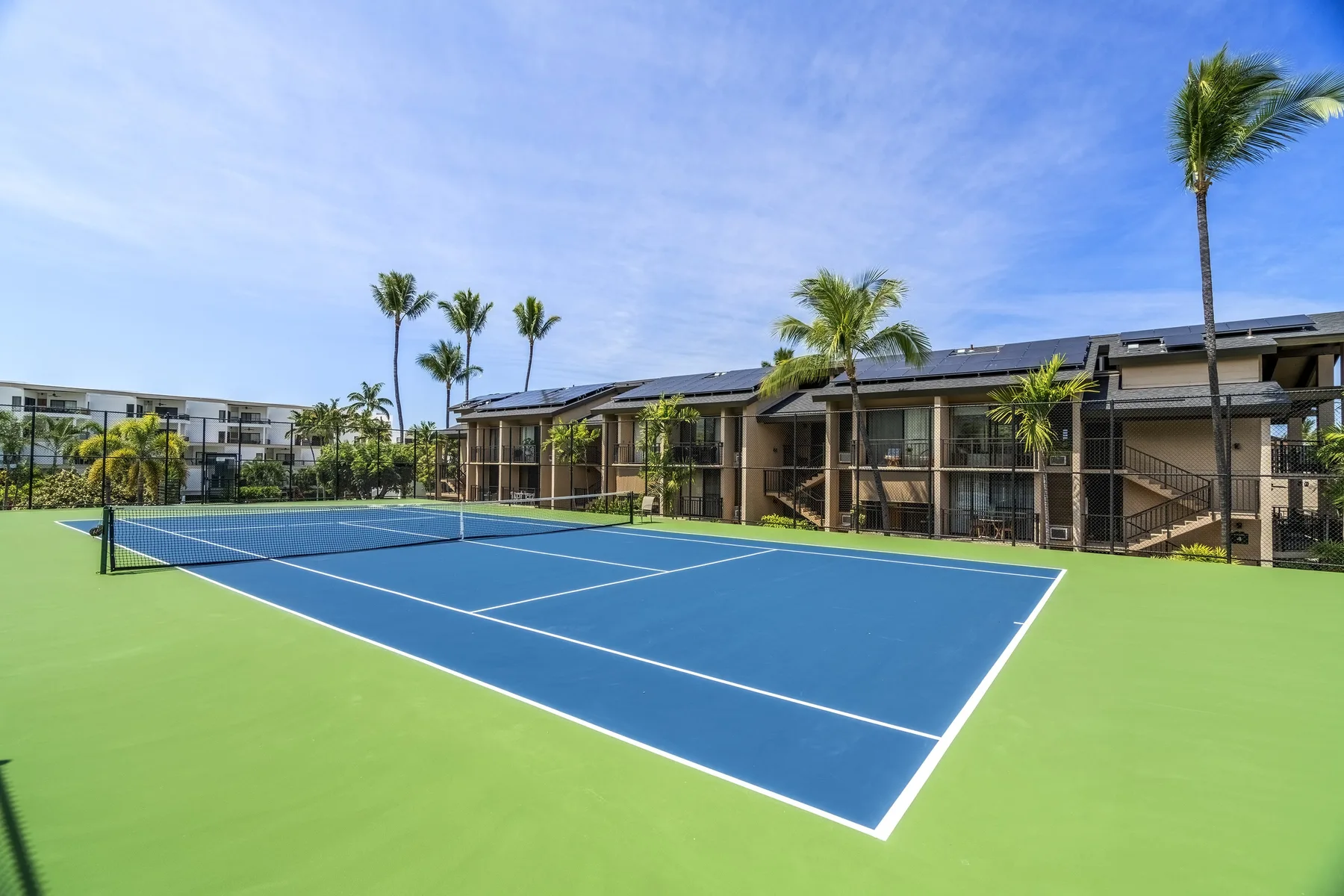 Una cancha de tenis al aire libre está junto a edificios bajos y palmeras.