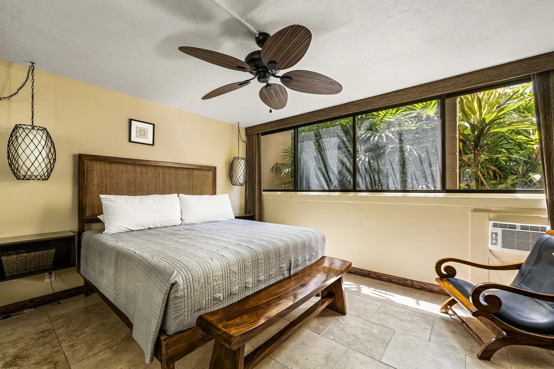 Un dormitorio con una cama, un ventilador de techo y grandes ventanas con vegetación al otro lado.