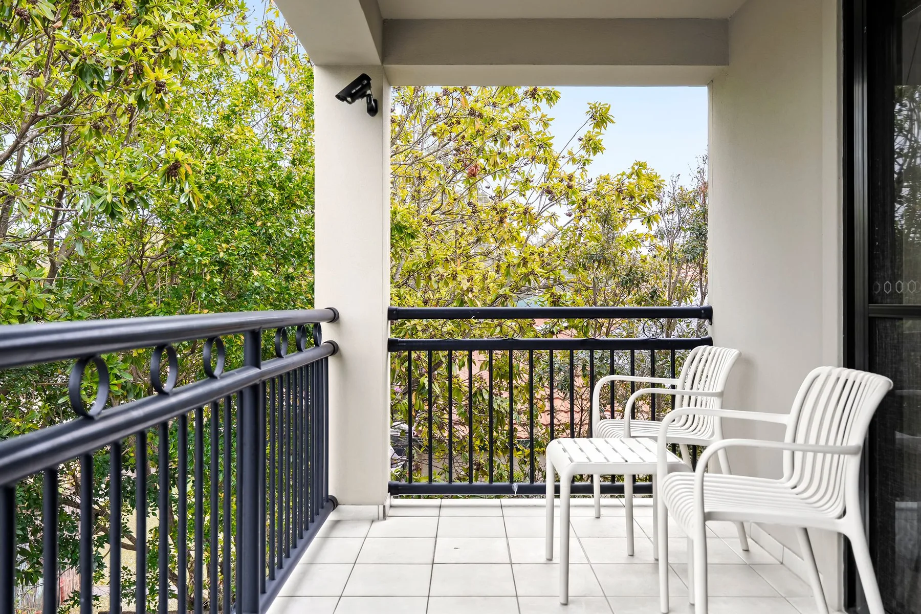 A covered balcony has white seating and black railings overlooking leafy trees.