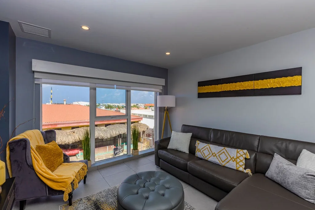 Your spacious living room features contemporary furnishings and art, with floor-to-ceiling windows framing colorful rooftops, a thatched palapa, and the vibrant neighborhood beyond.