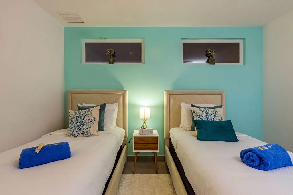 Step into your calming bedroom with two plush twin beds, crisp white linens, and vibrant blue towels welcoming you. The soothing turquoise accent wall sets a serene coastal mood for restful nights.