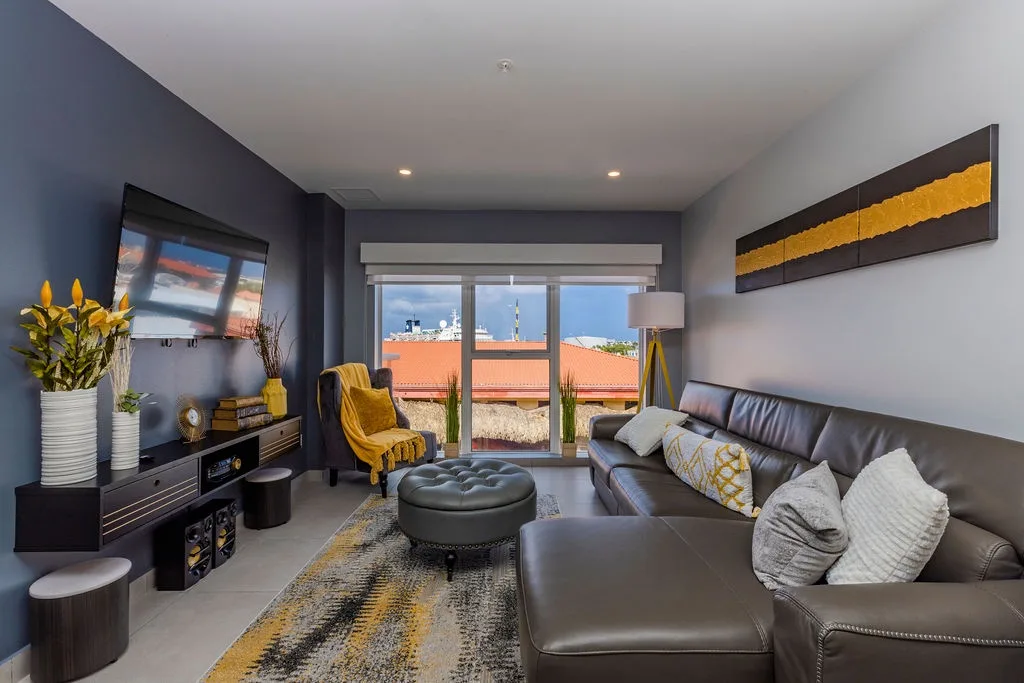 Sink into stylish comfort in your modern living space with rich leather seating, warm yellow accents, and natural light streaming through sliding glass doors to your private balcony.