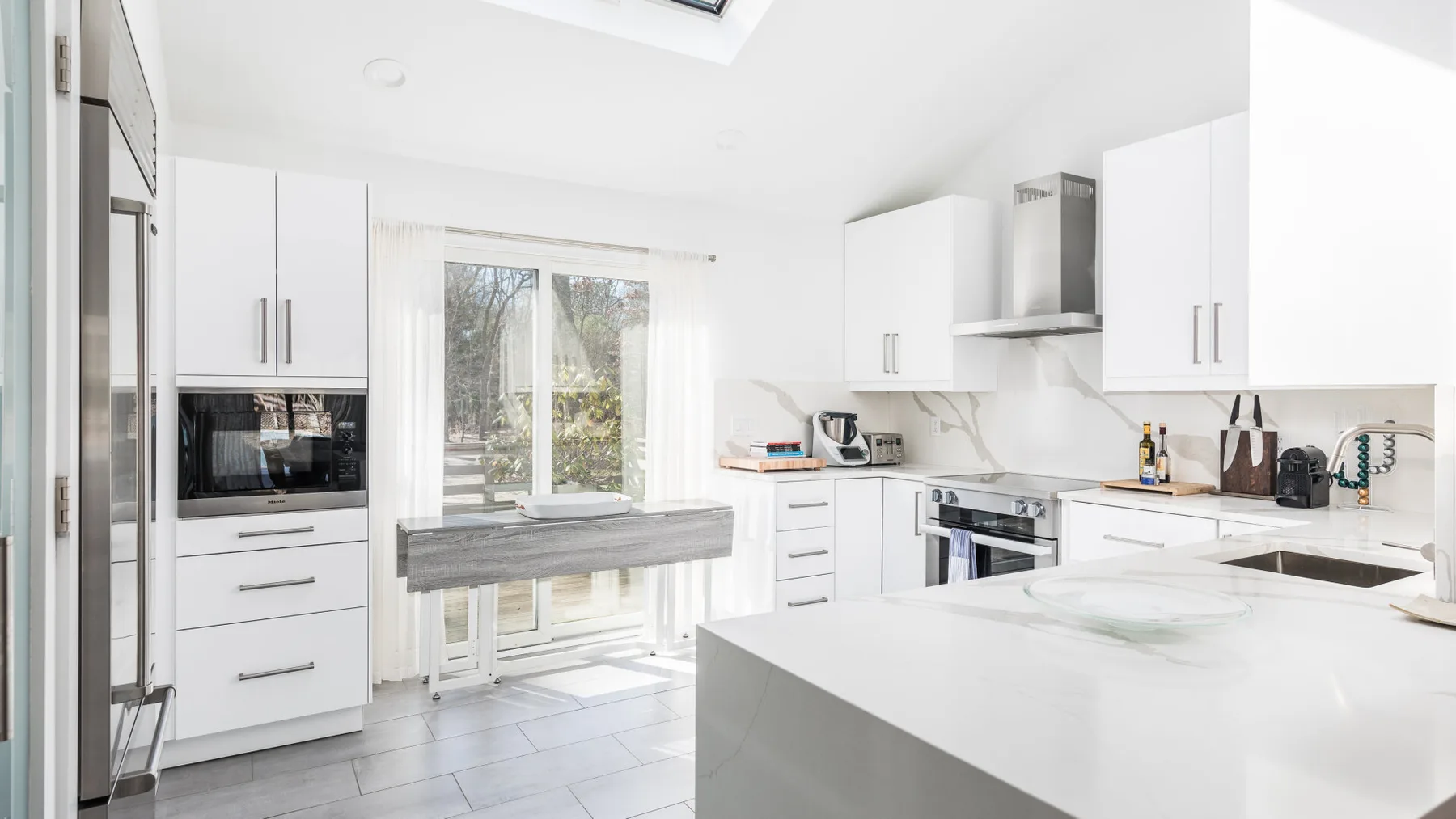 Una cocina blanca y luminosa con una isla grande y una ventana con vistas a la vegetación.