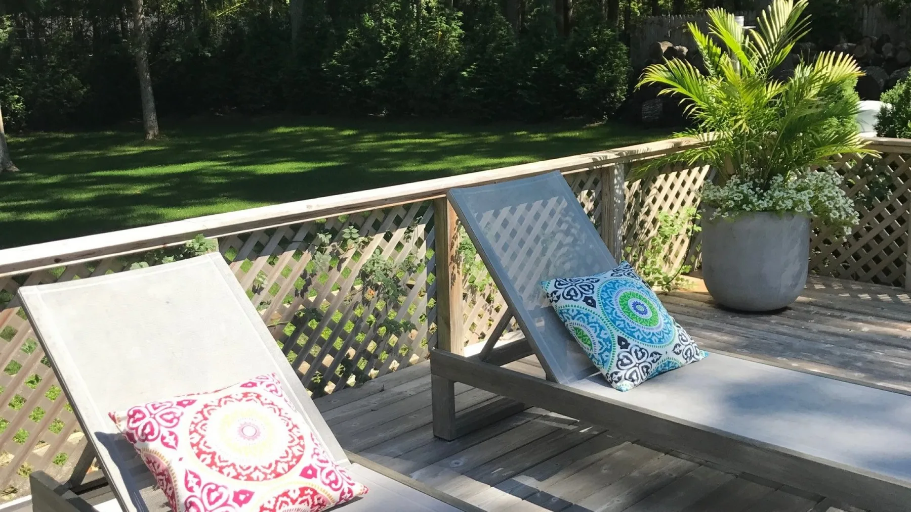 Una terraza de madera tiene dos tumbonas reclinables junto a una barandilla de celosía y un jardín.