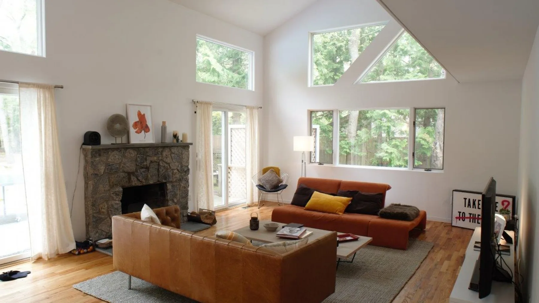 Una sala luminosa con chimenea de piedra, asientos y grandes ventanas con vista a los árboles.