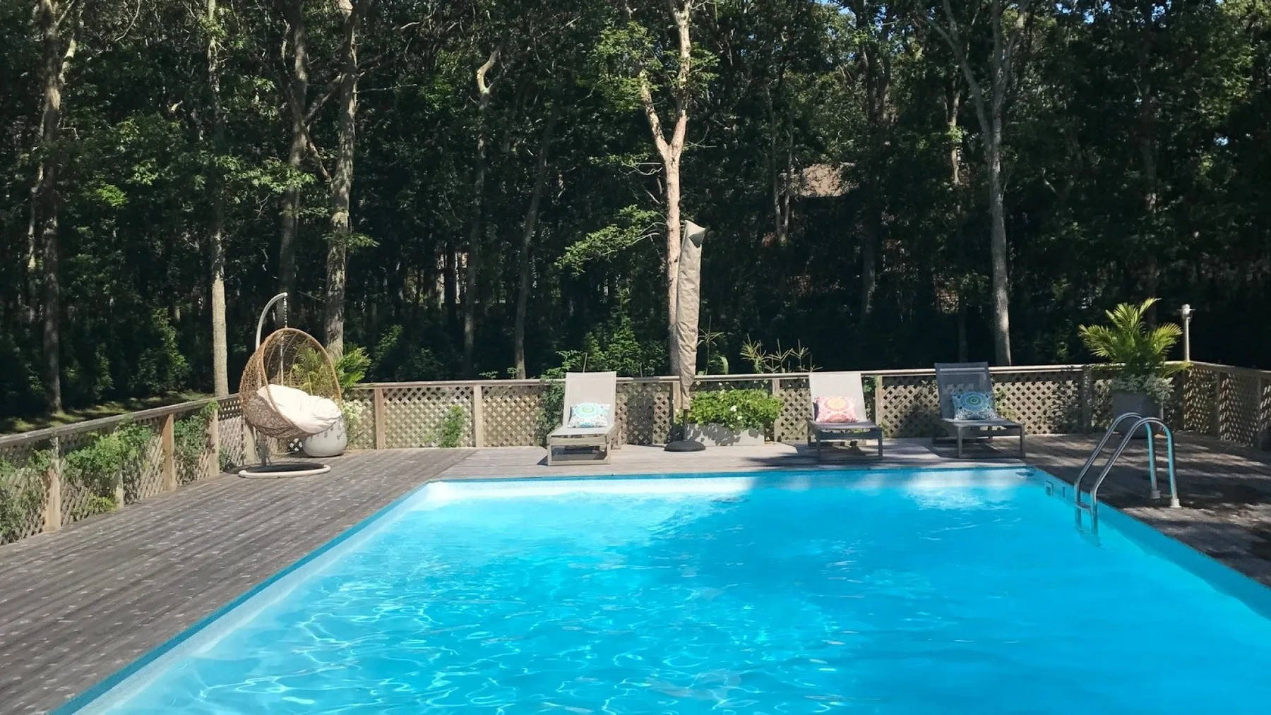 Una piscina al aire libre está junto a una terraza de madera, con árboles al otro lado de la barandilla.