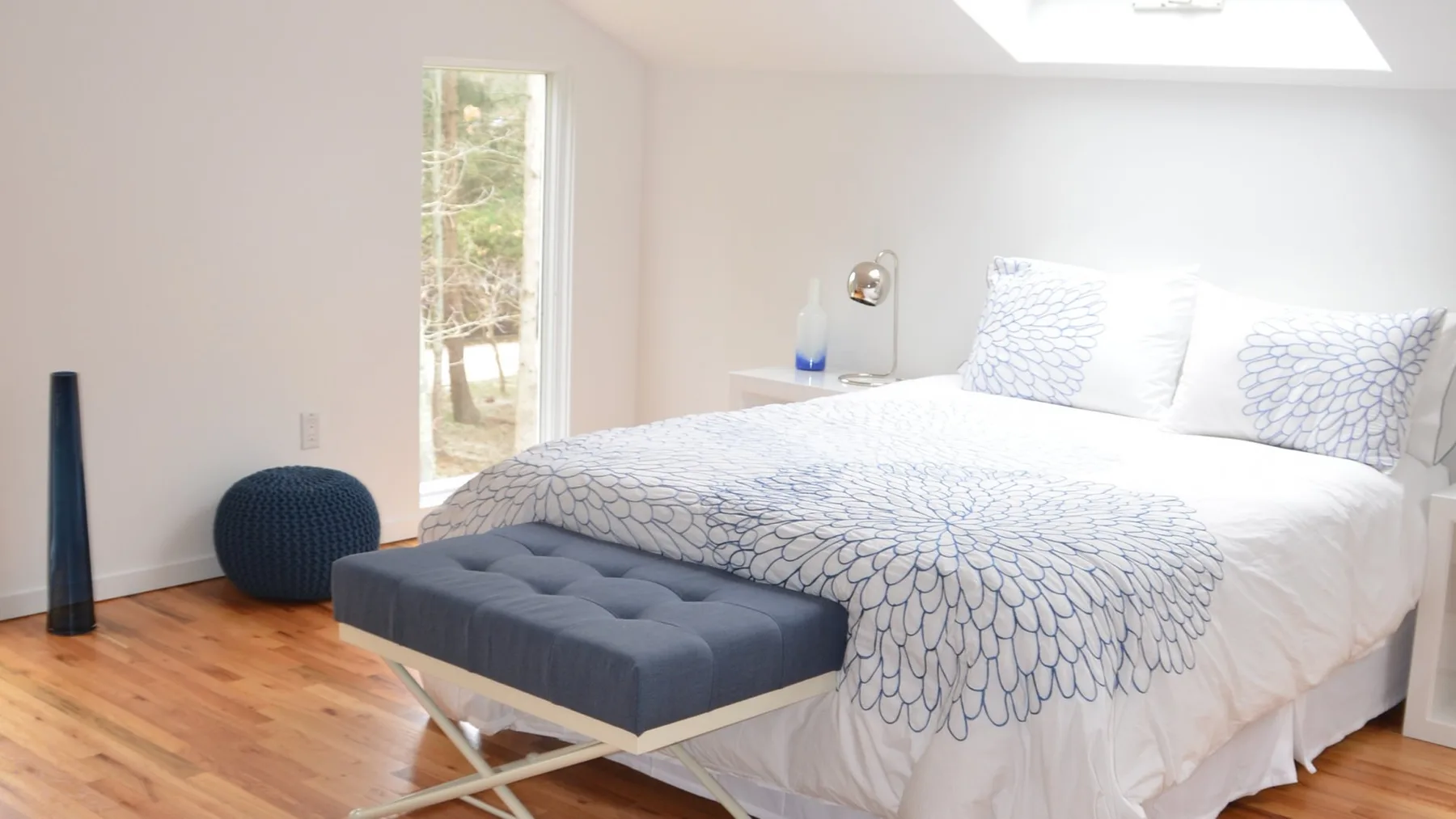 Un dormitorio luminoso con una cama vestida con ropa de cama blanca estampada y un banco acolchado.