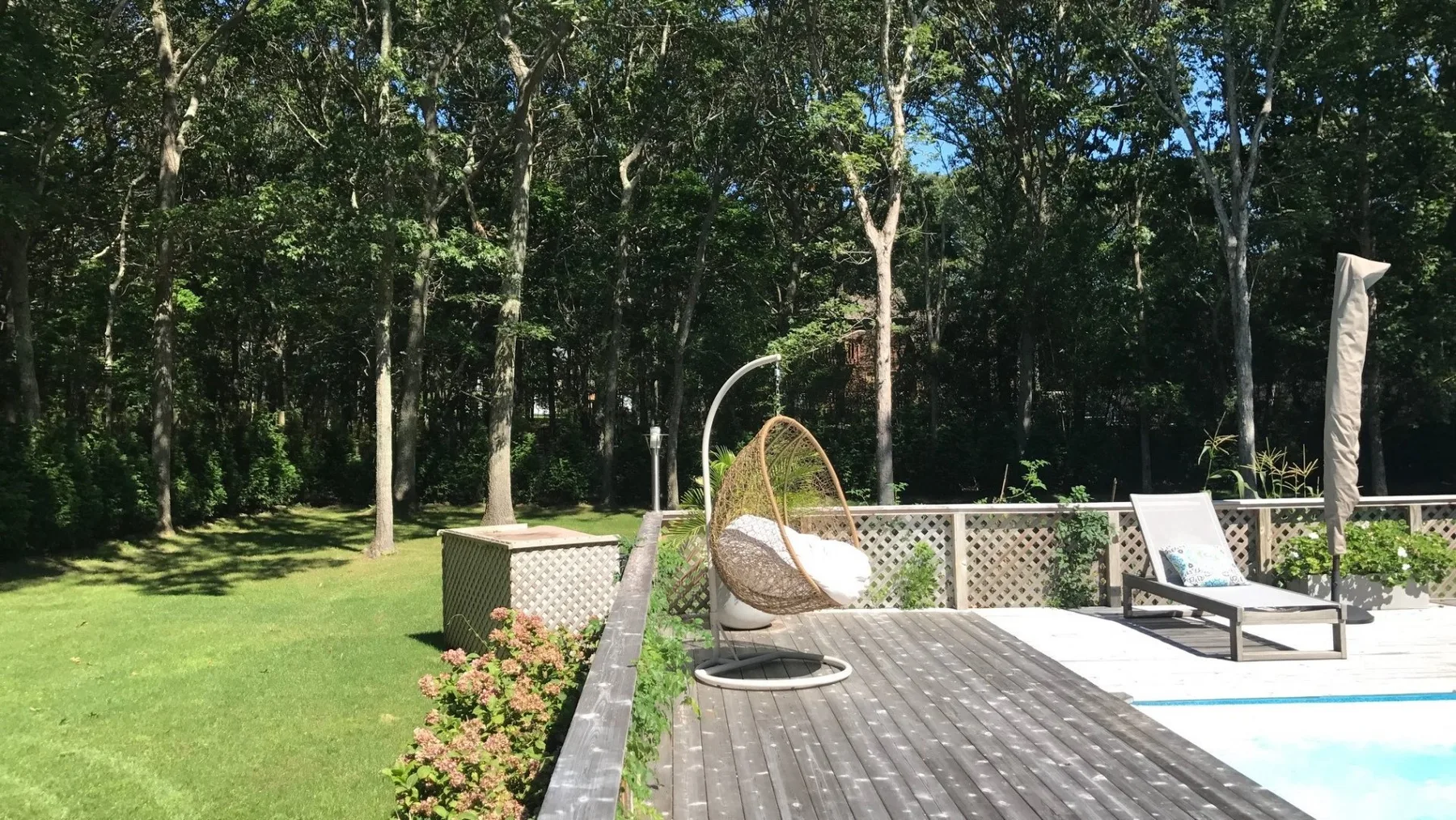 Una terraza soleada junto a una piscina, con una silla colgante y árboles alrededor.