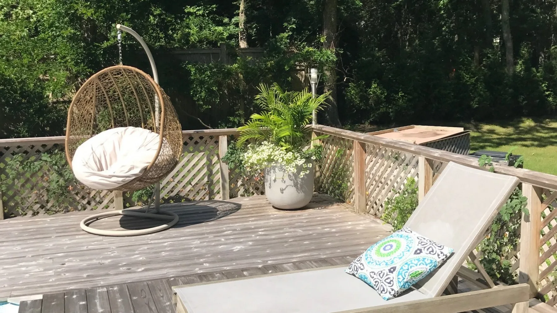 Una terraza exterior de madera tiene un asiento colgante, una silla reclinable y plantas en macetas.