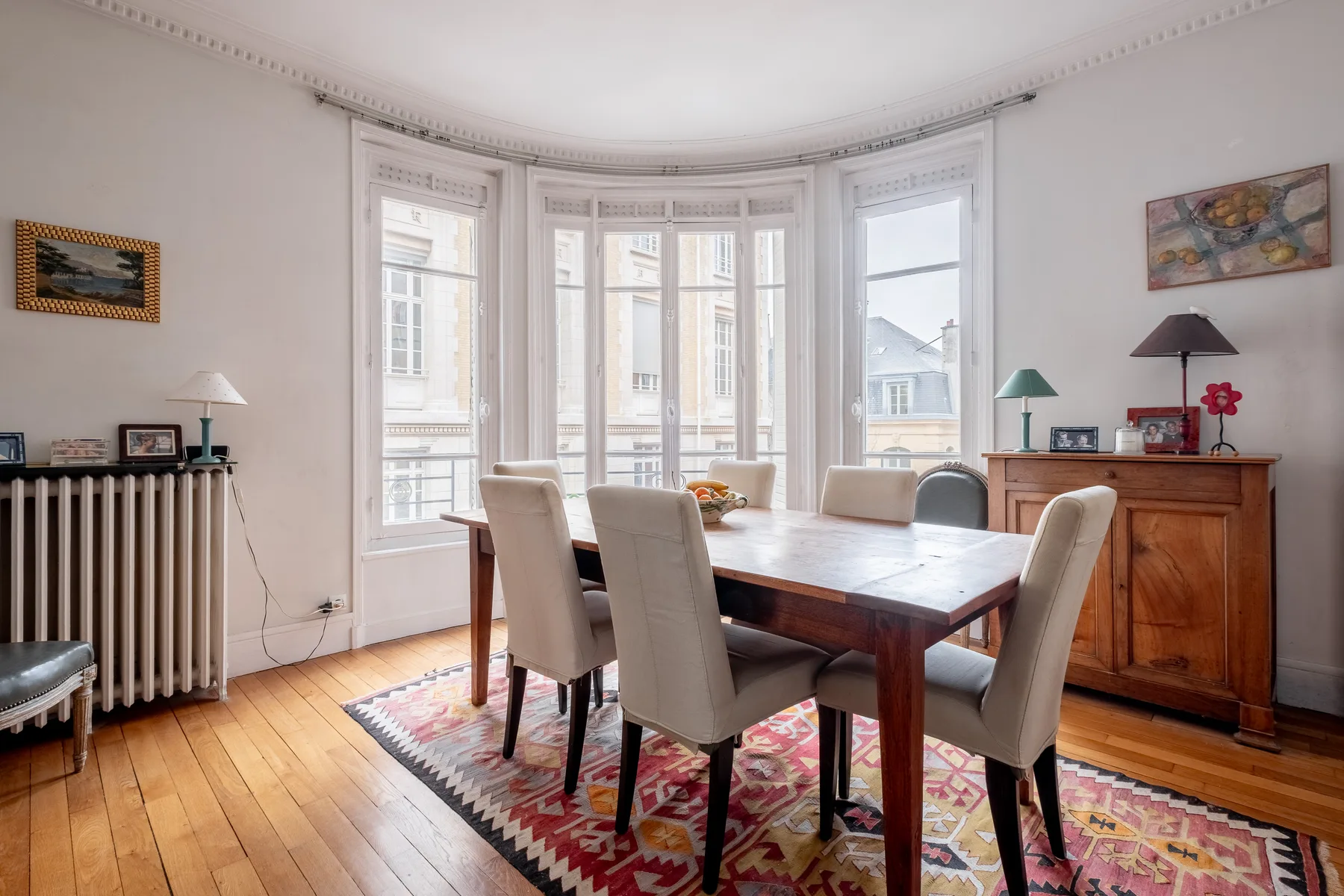 Comedor con mesa de madera, asientos tapizados y altos ventanales en mirador.