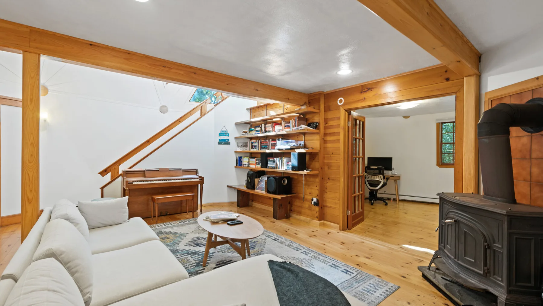 Una sala de estar con vigas de madera, asientos blancos, un piano y una estufa independiente.