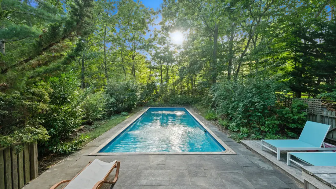 Una piscina está en una zona ajardinada pavimentada, rodeada de árboles y vegetación.