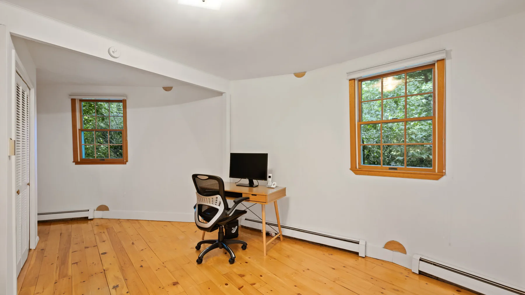 Una habitación luminosa con un escritorio y una silla entre dos ventanas.