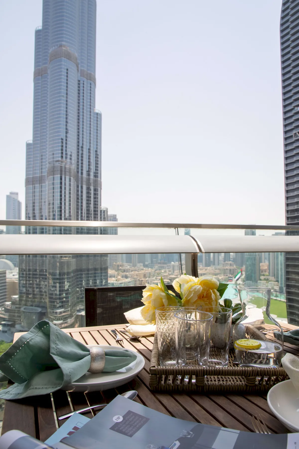 A balcony dining table overlooks a city skyline, with flowers and tableware.