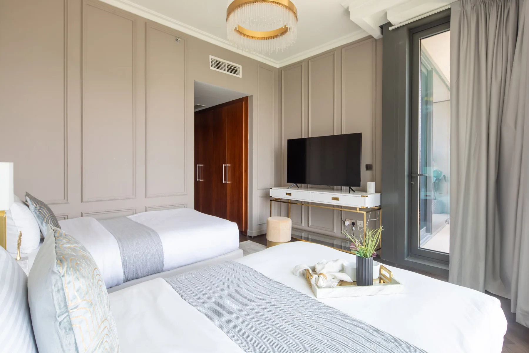A bright bedroom with two beds, a television, and a large curtained window.