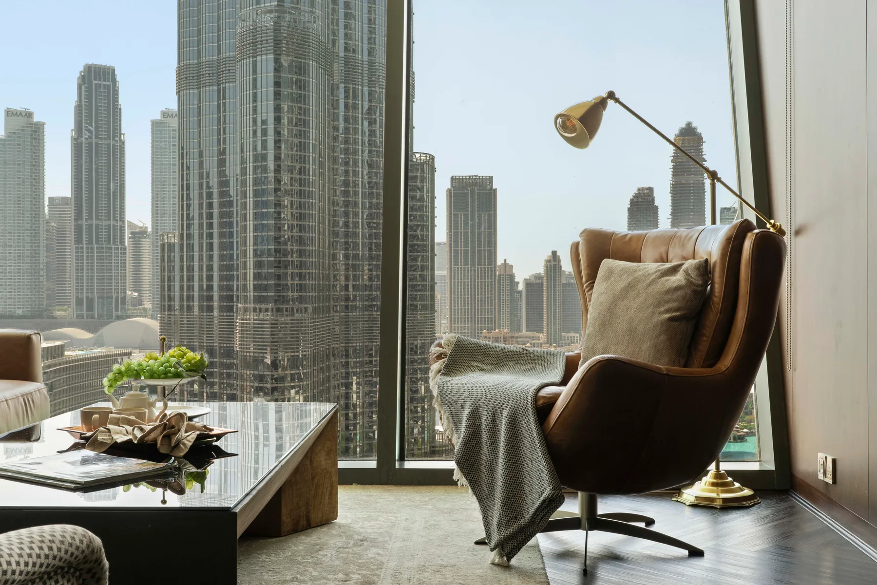 A modern living room with a lounge chair beside floor-to-ceiling windows overlooking city buildings.