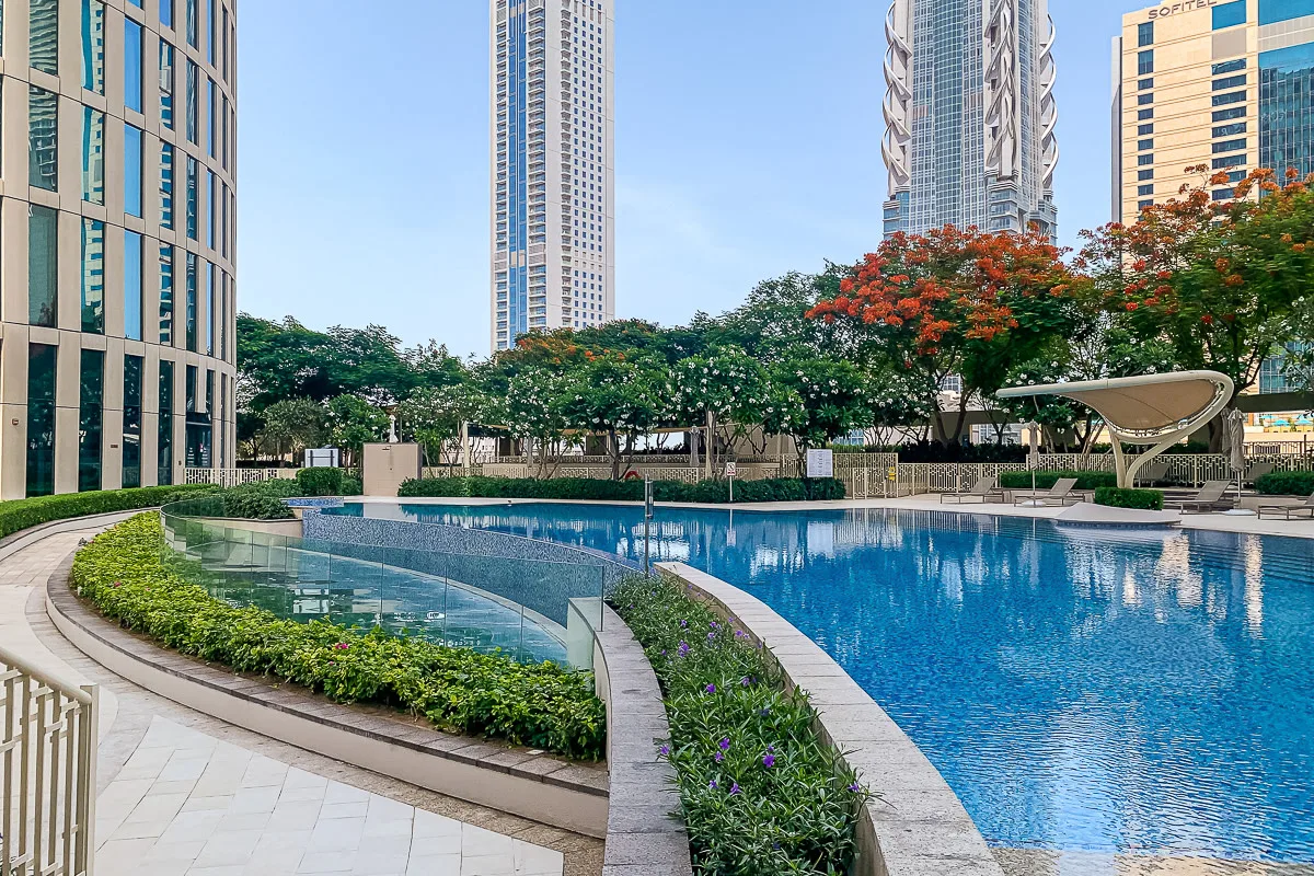 A landscaped outdoor swimming pool curves through a courtyard surrounded by trees and high-rise buildings.