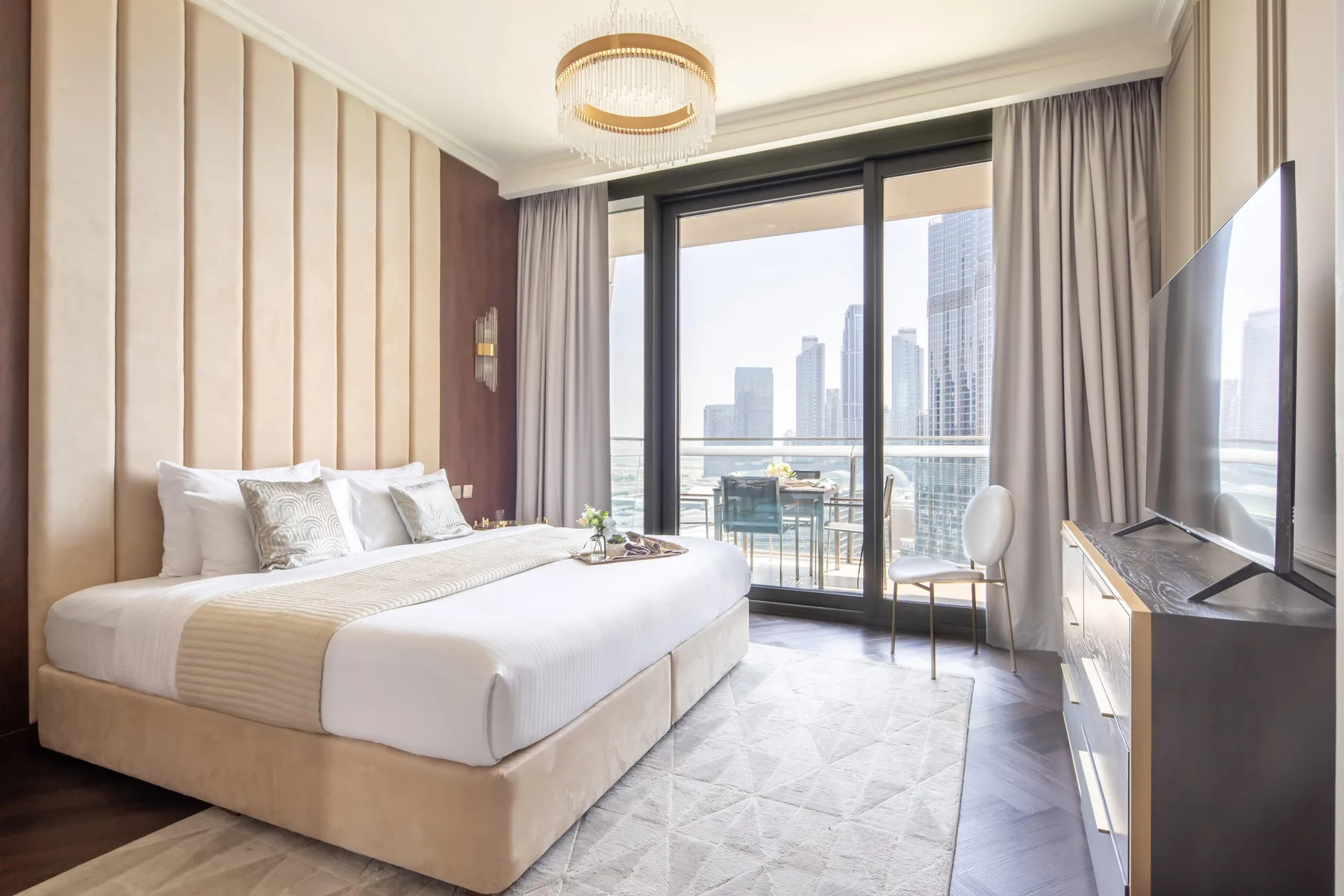 A bedroom with a neatly made bed beside floor-to-ceiling windows overlooking city buildings.