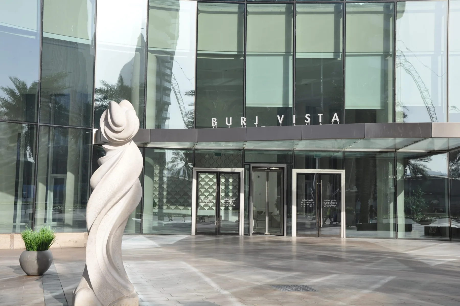 A glass-fronted building entrance faces a paved plaza with a sculptural feature.
