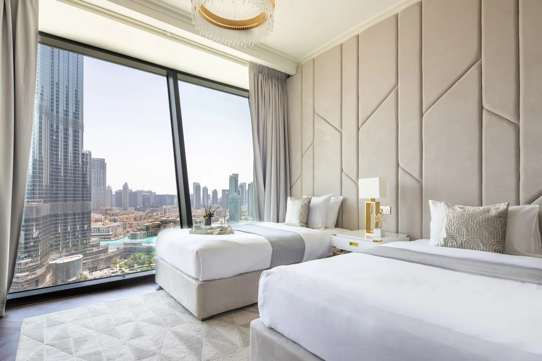 A bright bedroom with two beds beside a large window overlooking city buildings.