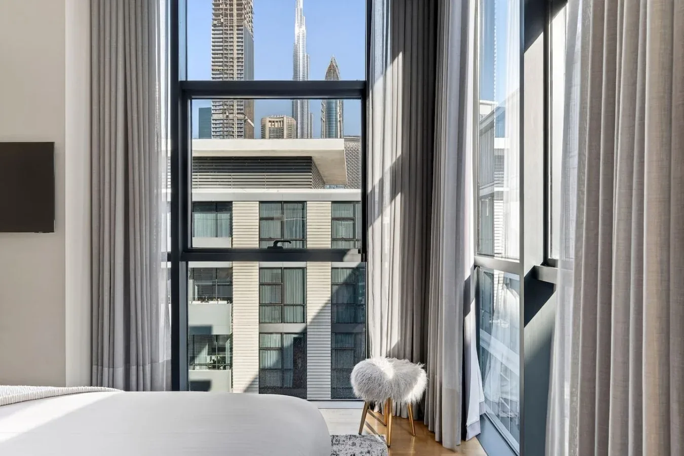 Un dormitorio con una cama junto a ventanas altas con vistas a edificios cercanos.