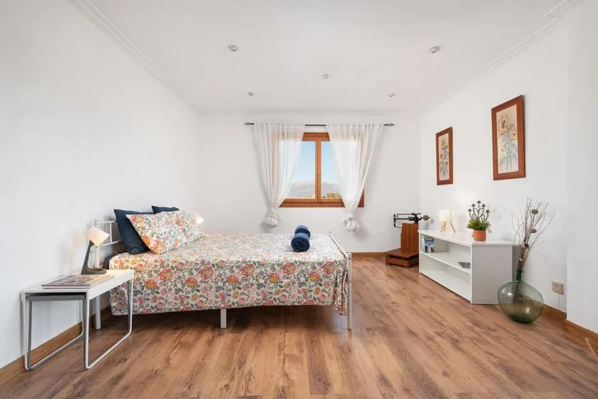 A bright bedroom has a floral bed, a window, and a white shelving unit.