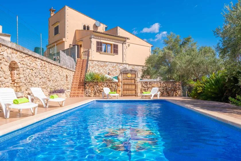 An outdoor swimming pool sits below a peach-colored house and stone retaining wall.