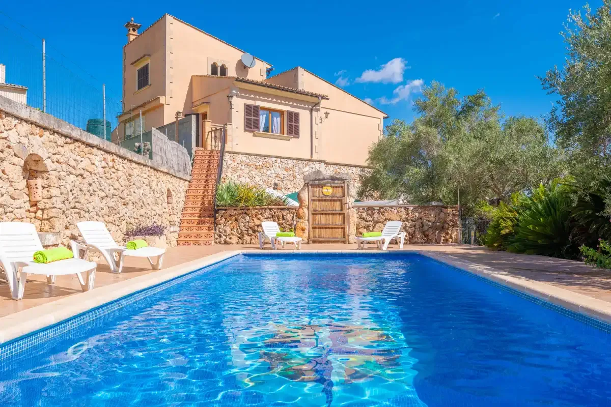 An outdoor swimming pool sits beside a stone wall and a peach-colored house.