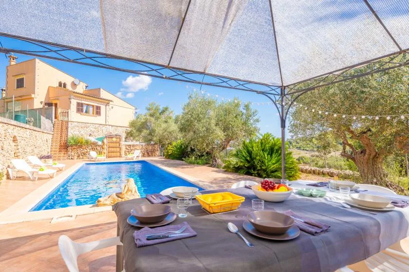 A shaded outdoor dining area beside a swimming pool and garden.