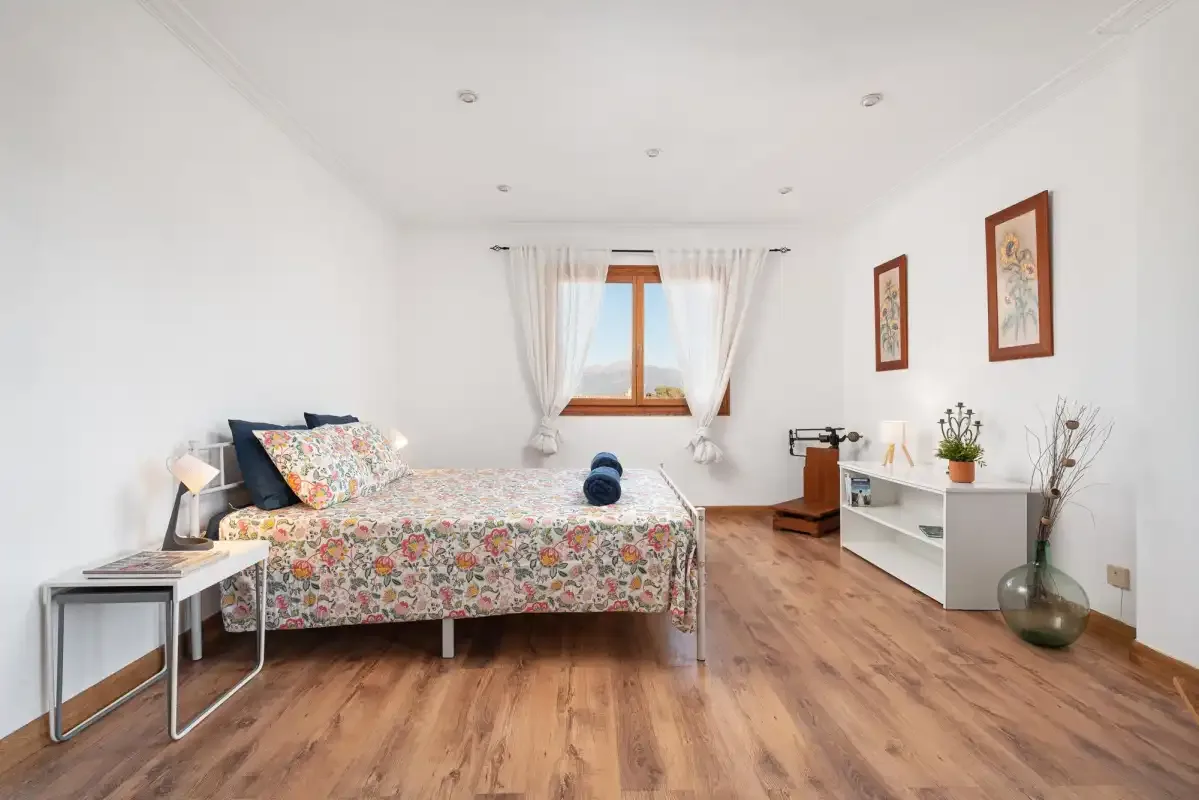 A bright bedroom with a floral bedspread and a window with sheer curtains.