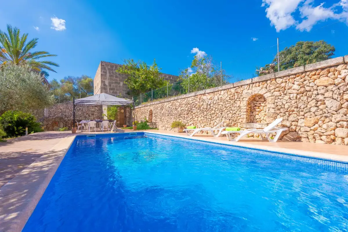 A swimming pool beside a stone wall, with lounge chairs and a shaded seating area.
