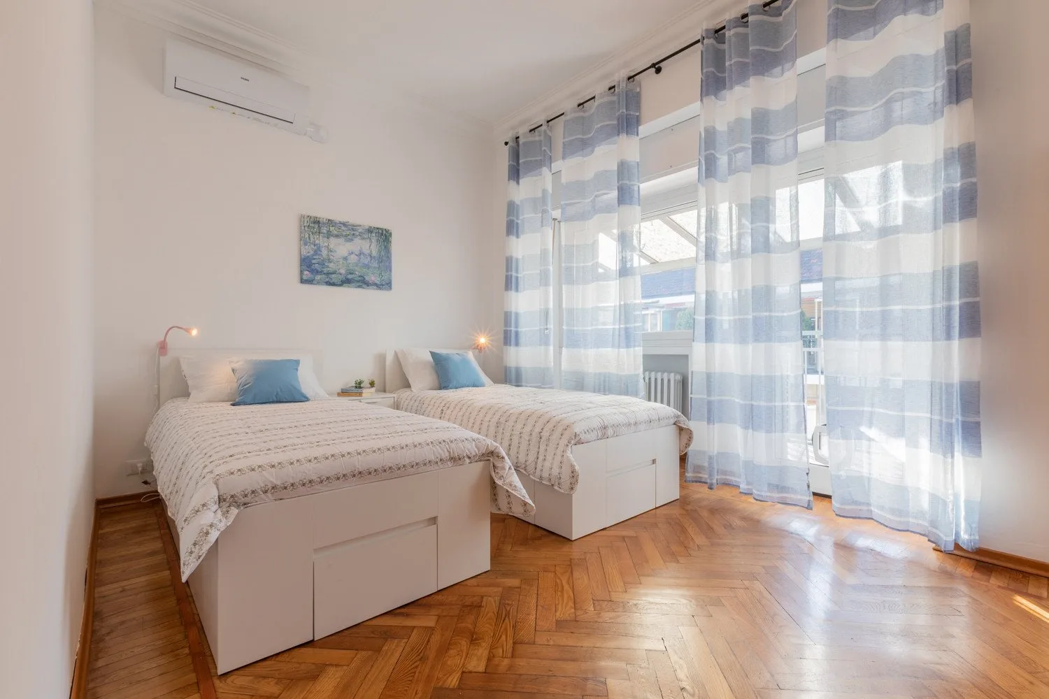A bright bedroom with two beds and blue curtains across the windows.