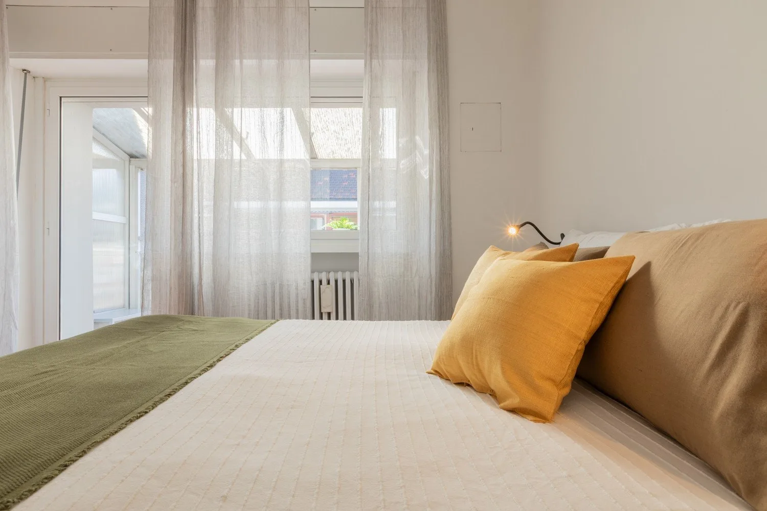 A bedroom with a neatly made bed, mustard-colored pillows, and sheer curtains.
